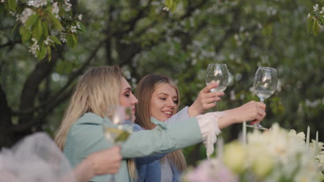 merry-hen-party-in-blooming-garden-women-are-clinking-glasses-and-laughing-sitting-at-table-in-orchard